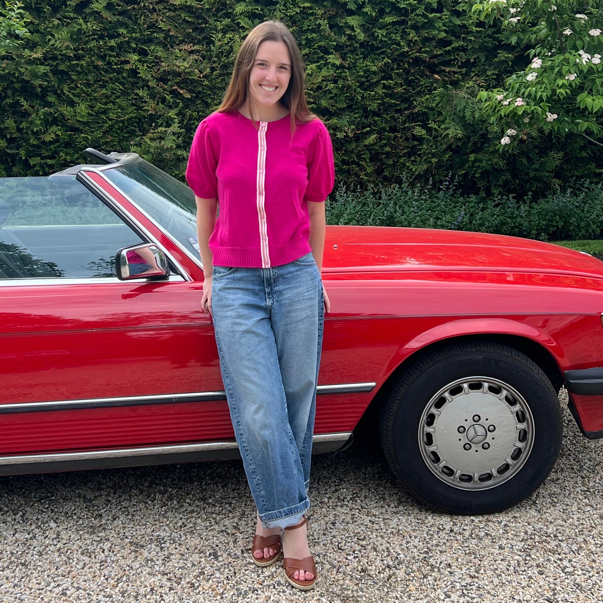 Woman standing next to a red convertible car in a driveway with greenery in the background wearing Noyac Knits Grace short puff-sleeve cardigan in gorgeous dark pink with signature ribbon trim knit from premium merino and styled with jeans and wedges