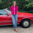 Woman standing next to a red convertible car in a driveway with greenery in the background wearing Noyac Knits Grace short puff-sleeve cardigan in gorgeous dark pink with signature ribbon trim knit from premium merino and styled with jeans and wedges