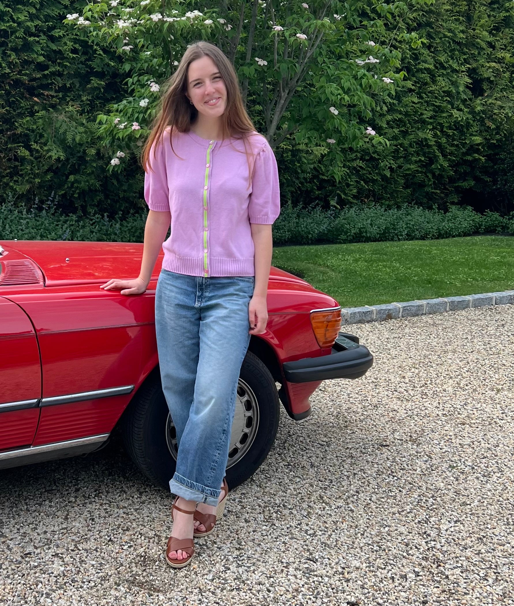 Woman in a Noyac Knits puff sleeve ribbon cardigan in premium lightweight merino styled with barrel jeans and wedges standing next to a red car on a gravel driveway.