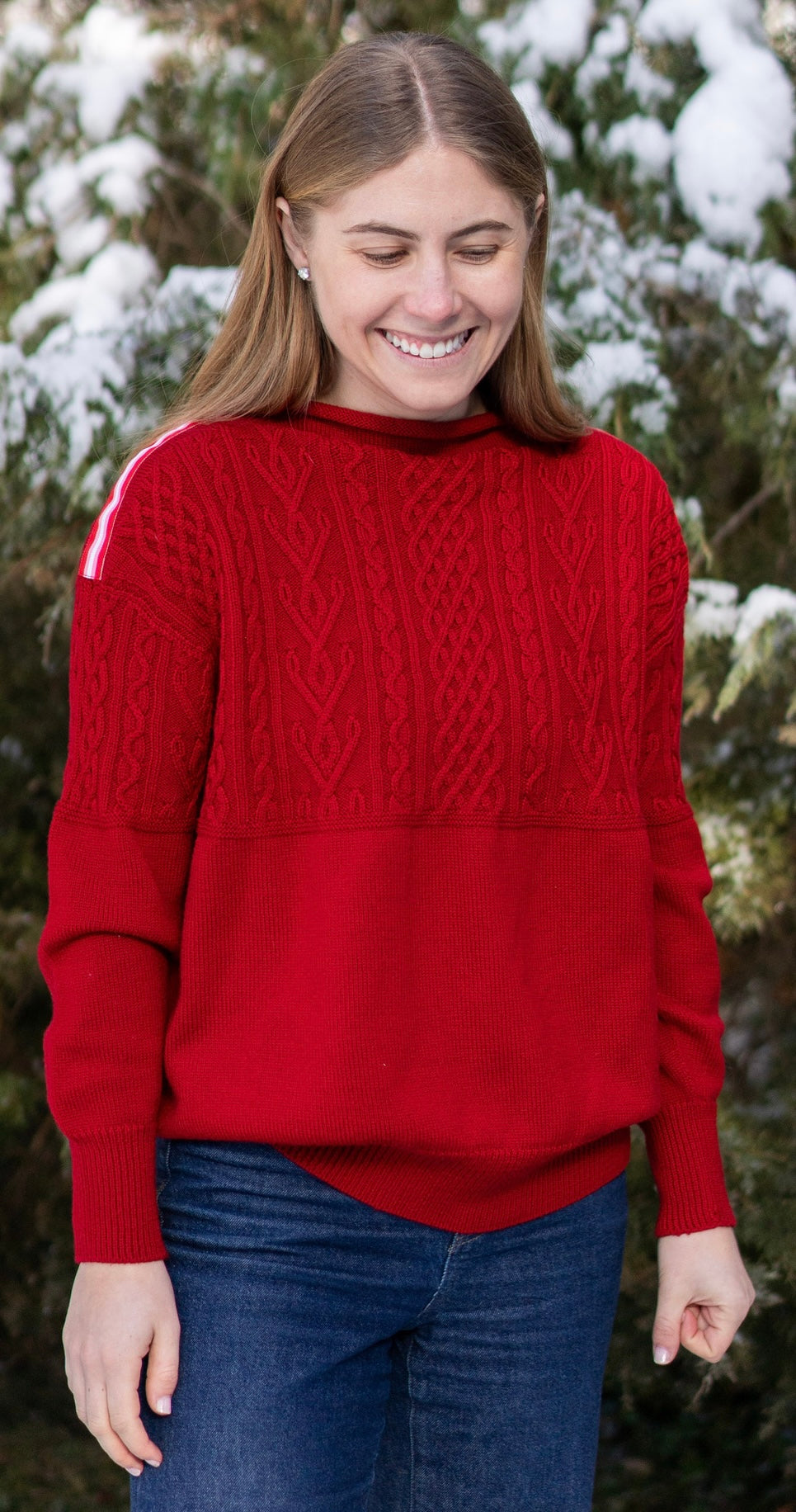 Woman wearing Noyac Knits Rhoda boatneck pullover sweater with beautiful cabling and signature red, pink and white striped ribbon trim on shoulders, knit in ultra soft premium merino wool, and styled with jeans in outdoor setting with snow covered trees in background.