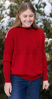 Woman wearing Noyac Knits Rhoda boatneck pullover sweater with beautiful cabling and signature red, pink and white striped ribbon trim on shoulders, knit in ultra soft premium merino wool, and styled with jeans in outdoor setting with snow covered trees in background.