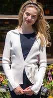 Woman wearing a Noyac Knits white wrap cardigan in environmentally sustainable ecoviscose worn over a featherweight scoop neck ecoviscose top in navy top in an outdoor setting with plants and flowers.