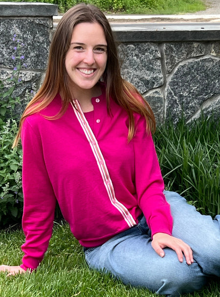 Woman in a Grace dark pink ribbon cardigan in lightweight all season merino shorts over matching crew neck shell sitting on grass with a stone wall and plants in the background