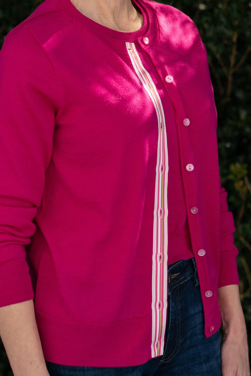 Woman wearing Noyac Knits Grace classic cardigan in beautiful raspberry pink with signature ribbon trim and pink buttons -- knit in lightweight premium merino -- styled with jeans and espadrilles in outdoor setting with greenery in background