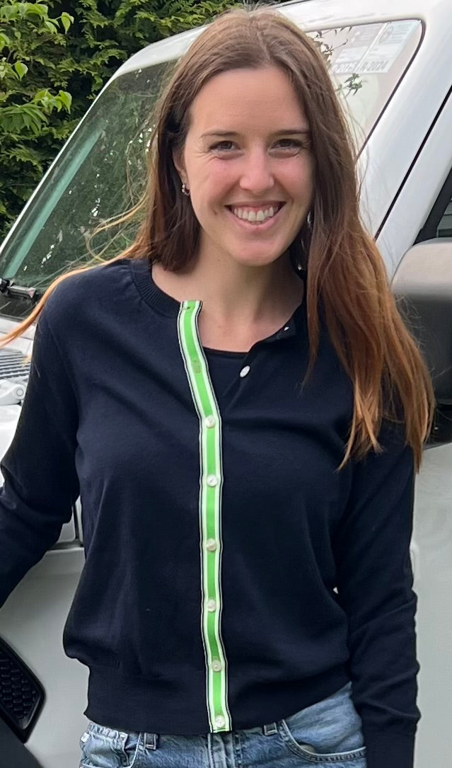 Woman wearing Noyac Knits navy Julie ribbon cardigan trimmed with with green, navy and white striped ribbon over a navy crew neck sleeveless sweater, both in premium Egyptian cotton, styled with  jeans standing in front of white jeep outside