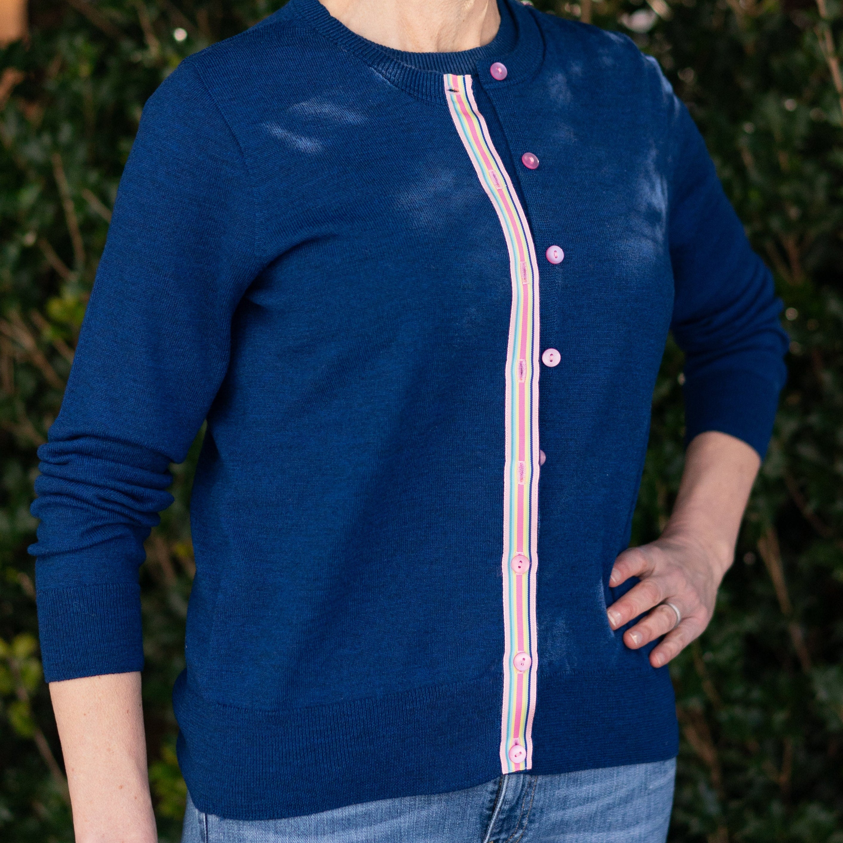Woman wearing a Noyac Knits Sissy classic cardigan in gorgeous cobalt blue with signature pink rainbow striped ribbon trim  and pink buttons made in preimium lightweight merino styled with jeans against a green leafy background