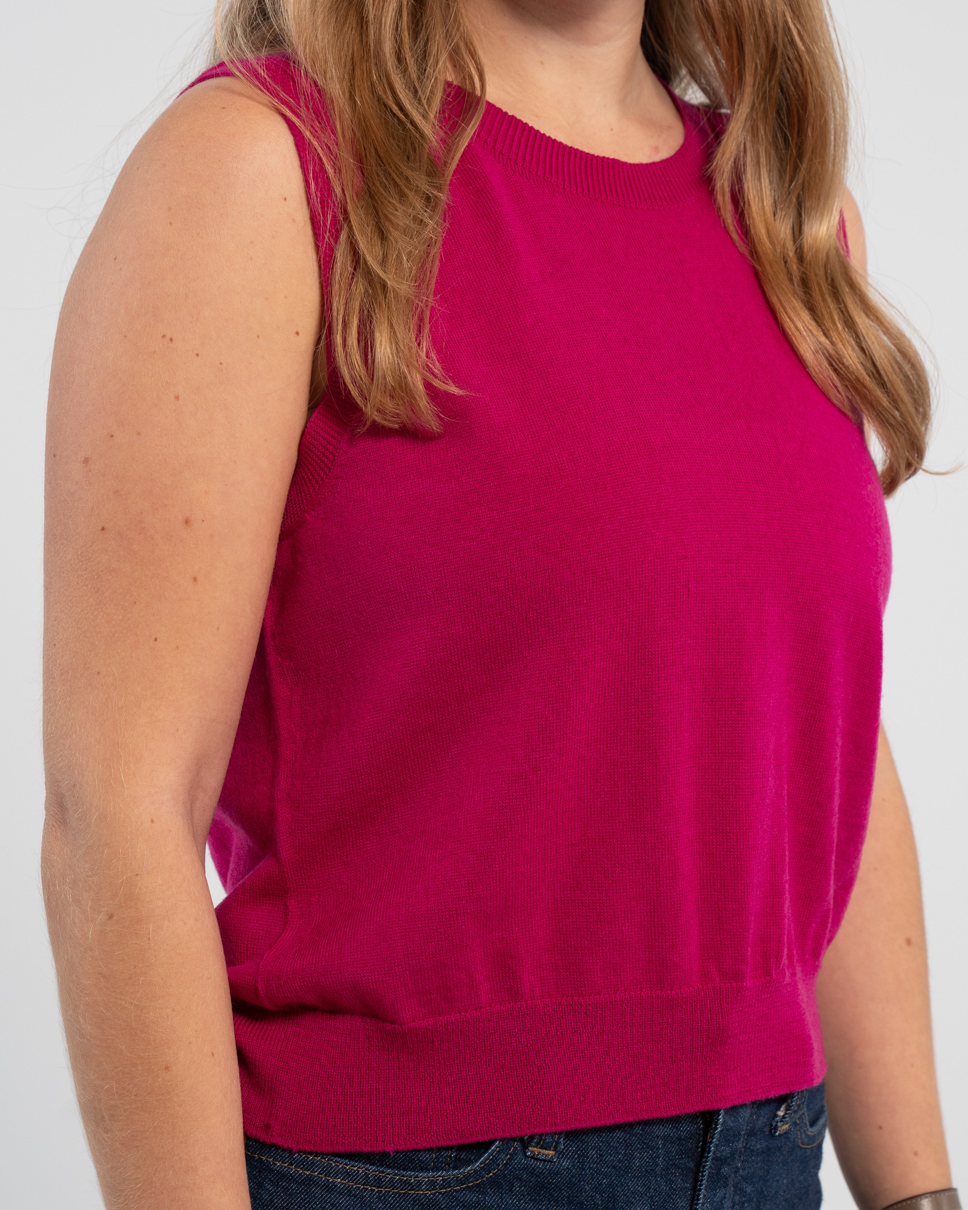 Close up on woman wearing Noyac Knits crop raspberry pink sleeveless sweater in ultrafine merino wool and jeans on plain background.