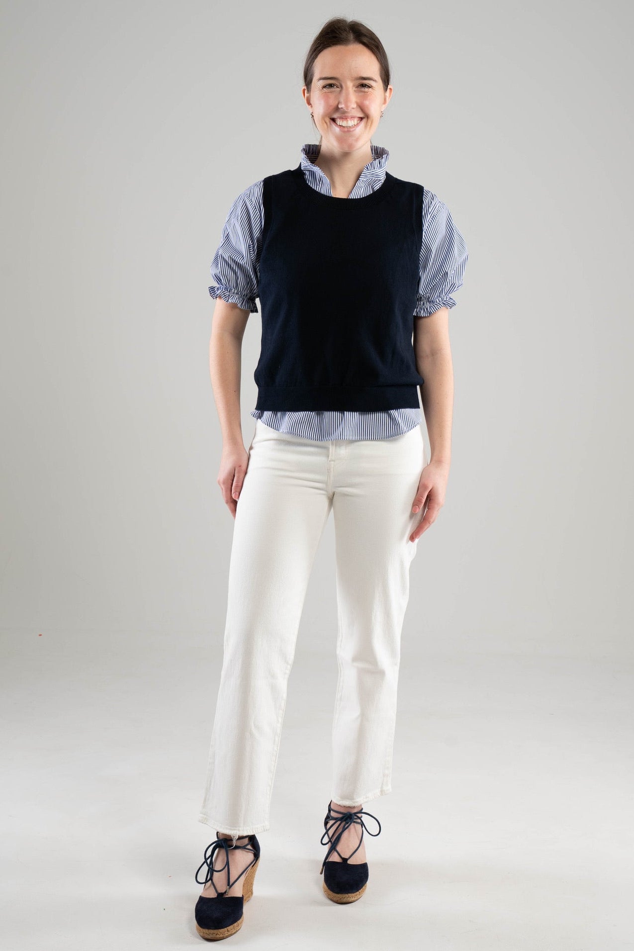 Woman wearing a Noyac Knits classic navy crew neck sleeveless shell styled over a blue and white puff-sleeve, ruffle neck blouse  with white pants and navy espadrilles on a white  background