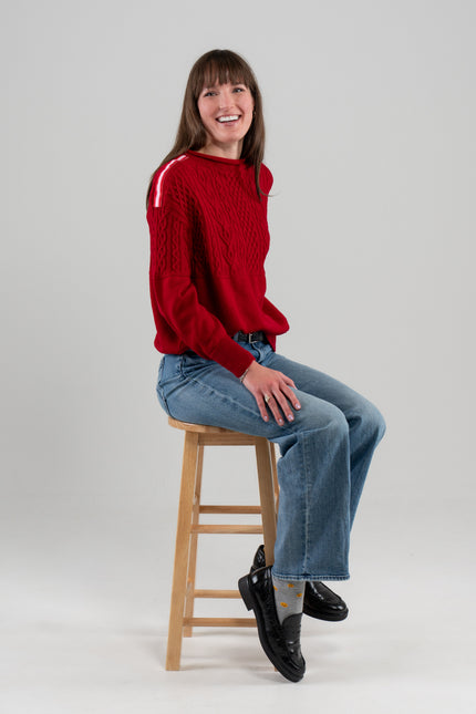 Woman wearing Noyac Knits Rhoda boatneck pullover sweater with beautiful cabling and signature red, pink and white striped ribbon trim on shoulders, knit in ultra soft premium merino wool, and styled with jeans and loafers seated on stool on plain background.