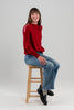 Woman wearing Noyac Knits Rhoda boatneck pullover sweater with beautiful cabling and signature red, pink and white striped ribbon trim on shoulders, knit in ultra soft premium merino wool, and styled with jeans and loafers seated on stool on plain background.