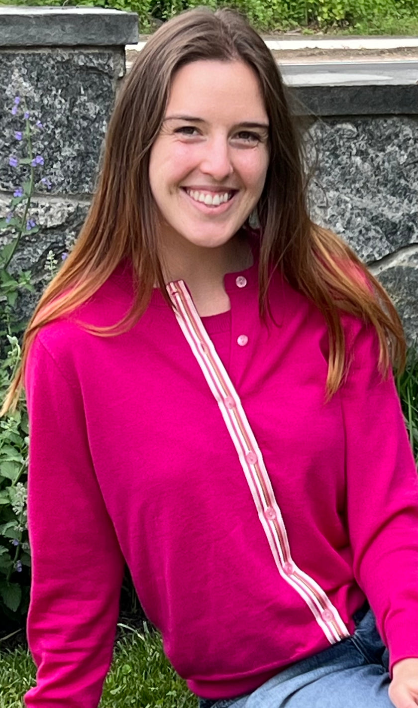 Woman wearing Noyac Knits Grace classic cardigan in beautiful raspberry pink with signature ribbon trim and pink buttons -- knit in lightweight premium merino -- styled with jeans in outdoor setting with greenery and stone wall in background
