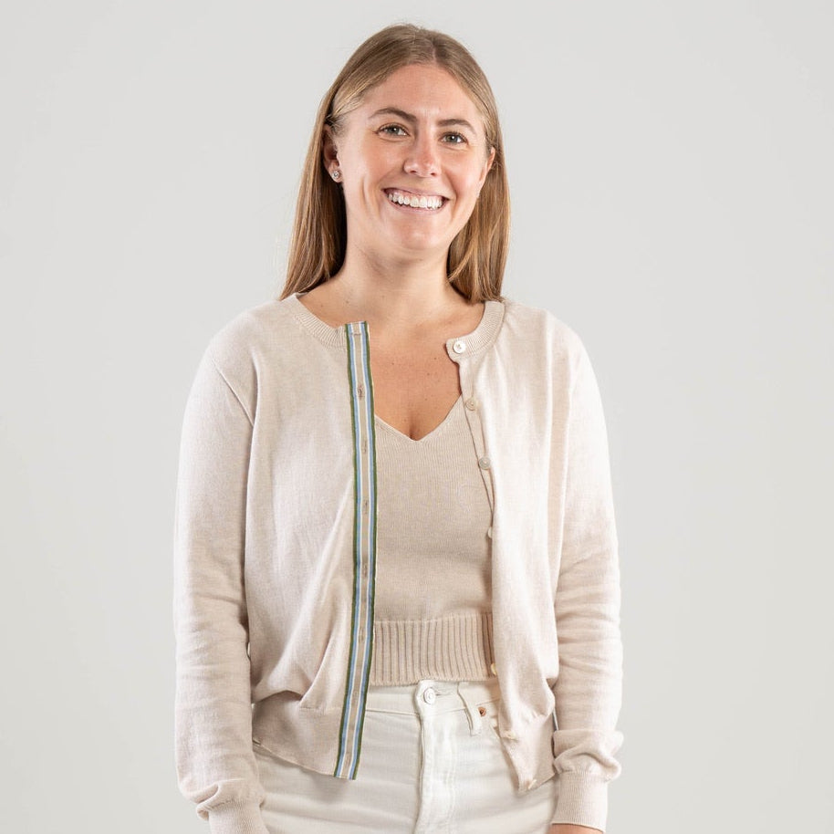 Woman wearing Noyac Knits Lynn classic cardigan in light tan  with army green, light blue, white, and tan striped ribbon trim and mother of pearl butons, in premium Egyptian cotton, styled with white jeans and white background 