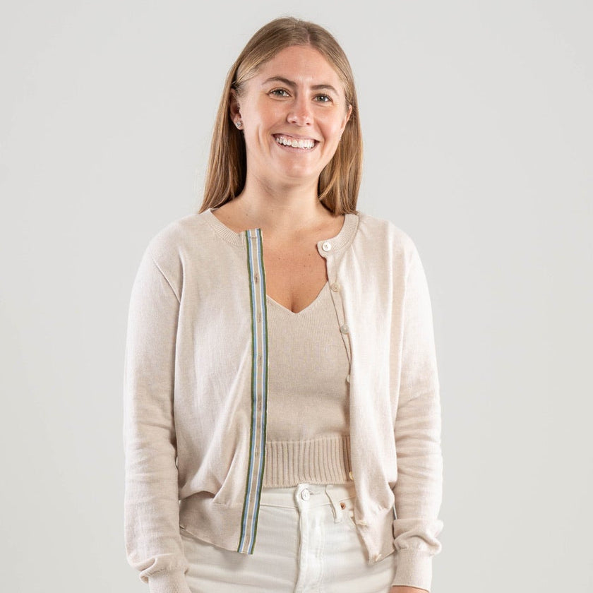 Woman wearing Noyac Knits Lynn classic cardigan in light tan  with army green, light blue, white, and tan striped ribbon trim and mother of pearl butons, in premium Egyptian cotton, styled with white jeans and white background 