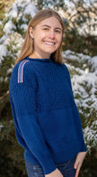 Woman wearing Noyac Knits cobalt blue boatneck pullover sweater with gorgeous cable detailing and signature ribbon trim in stripes of blue, pink and yellow on shoulder, knit in premium, soft merino wool  in outdoor setting with snow covered trees in the background.