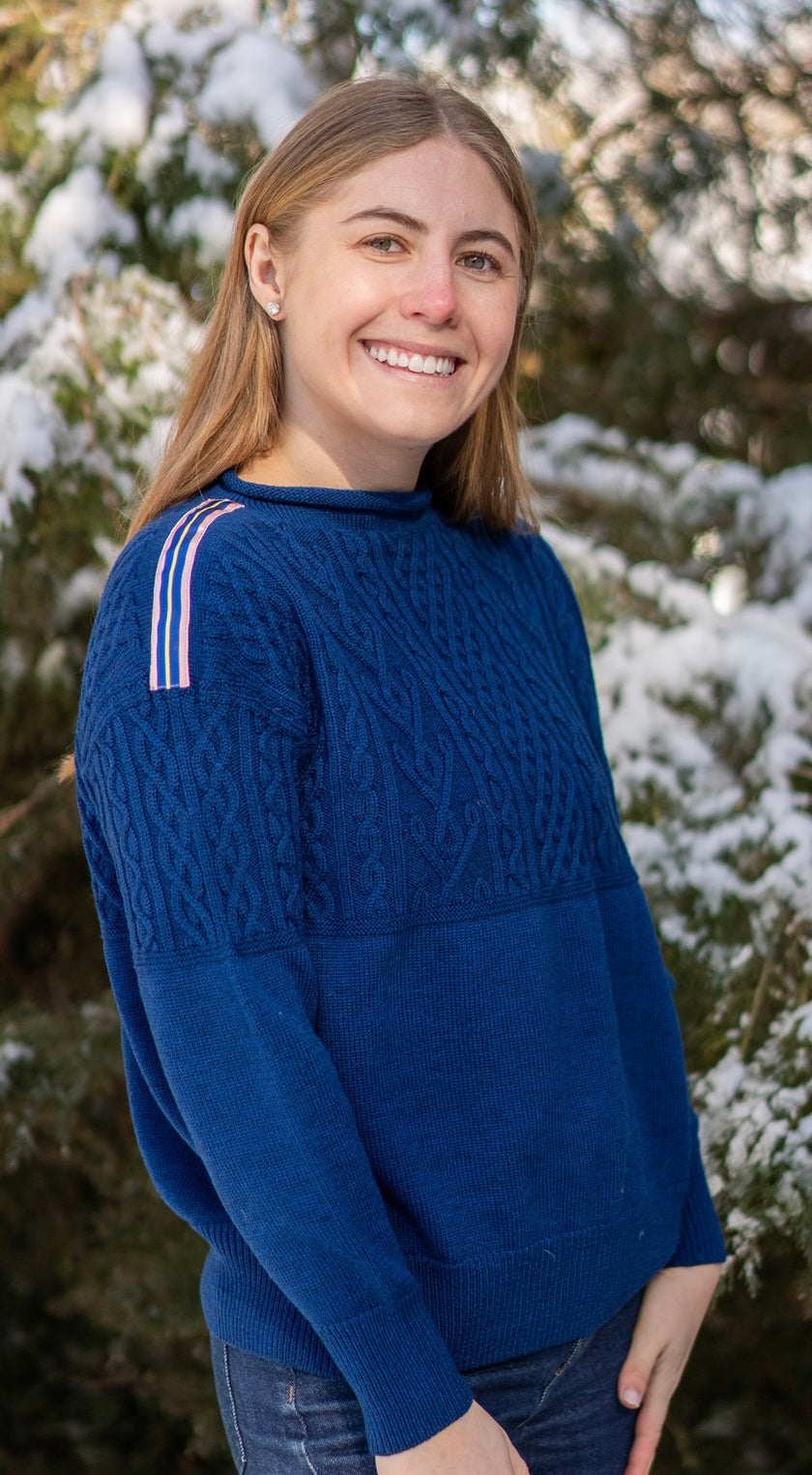 Woman wearing Noyac Knits cobalt blue boatneck pullover sweater with gorgeous cable detailing and signature ribbon trim in stripes of blue, pink and yellow on shoulder, knit in premium, soft merino wool  in outdoor setting with snow covered trees in the background.