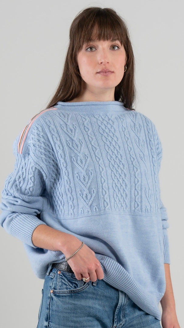 Woman wearing Noyac Knits Channing boatneck pullover in beautiful baby blue featuring cabling and signature ribbon trim on shoulder, knit in premium soft merino wool and styled with  jeans and loafers on plain background.