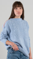 Woman wearing Noyac Knits Channing boatneck pullover in beautiful baby blue featuring cabling and signature ribbon trim on shoulder, knit in premium soft merino wool and styled with  jeans and loafers on plain background.