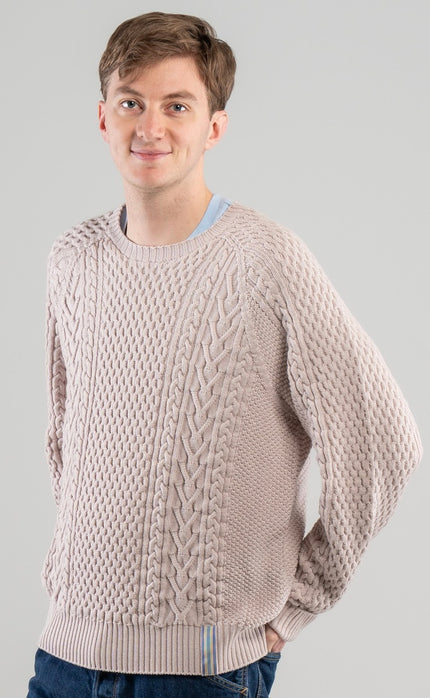 Man wearing Noyac Knits Cooper beige cable-knit sweater with signature ribbon trim on hem -- knit in premium merino wool -- styled with  jeans  on plain background.