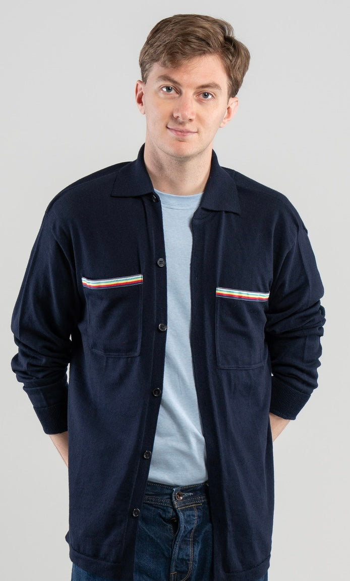 Man wearing Noyac Knits Finn knit overshirt in navy premium soft lightweight merino wool with signature ribbon detailing on pockets styled  over light blue t-shirt with jeans on plain background.