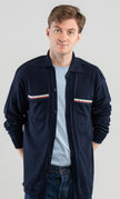 Man wearing Noyac Knits Finn knit overshirt in navy premium soft lightweight merino wool with signature ribbon detailing on pockets styled  over light blue t-shirt with jeans on plain background.