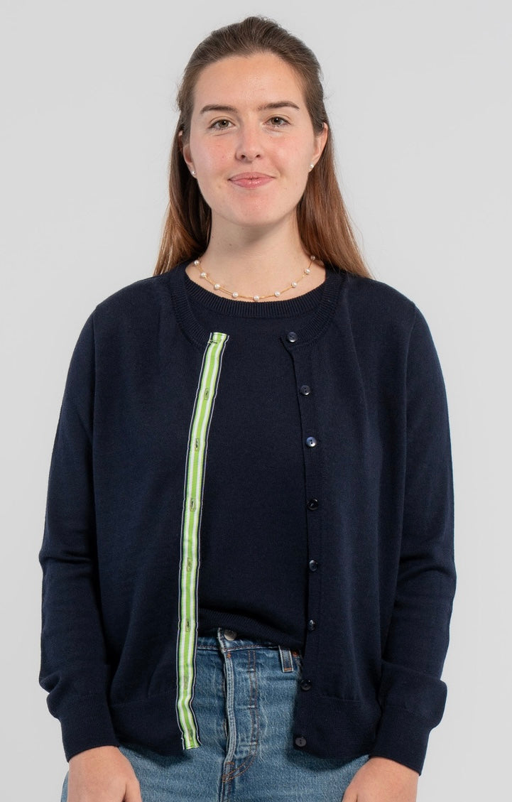 Woman wearing unbuttoned Noyac Knits Julie classic cardigan in navy trimmed with signature  navy buttons and green, white and navy grosgrain ribbon -- knit in premium, lightweight merino -- over matching navy shell sweater, also in soft merino and styled with jeans  on plain background.
