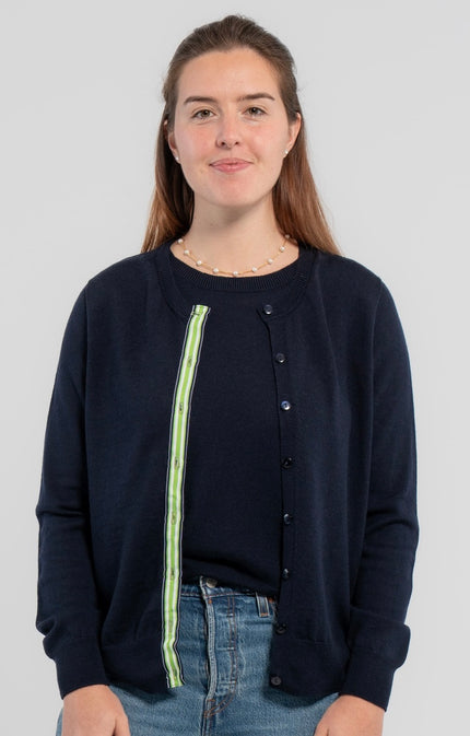Woman wearing unbuttoned Noyac Knits Julie classic cardigan in navy trimmed with signature  navy buttons and green, white and navy grosgrain ribbon -- knit in premium, lightweight merino -- over matching navy shell sweater, also in soft merino and styled with jeans  on plain background.