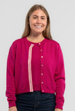 Woman wearing Noyac Knits Grace Cardigan in raspberry pink premium merino wool trimmed with signatrue  pink buttons and pink, white, green striped grosgrain ribbon trim and jeans on plain background.