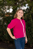 Woman wearing a dark pink Noyac Knits short puff sleeve ribbon cardigan trimmed with with pink, white, green striped ribbon and pink buttons --  knit is premium, lightweight merino -- styled with blue jeans in outdoor setting with greenery in the background