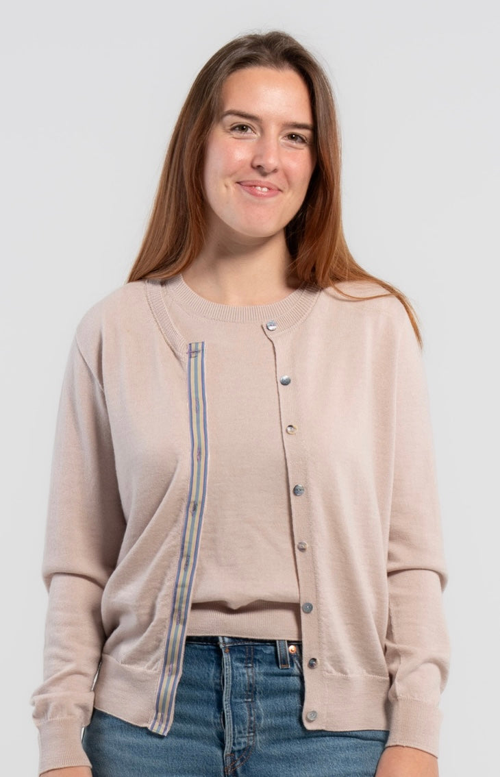 Woman wearing Noyac Knits Isabelle Classic Cardigan in light beige trimmed with signature grosgrain ribbon in stripes of tan, purple, lavender, and teal and Japanese mussel shell buttons -- knit in lightweight premium merino -- styled with matching merino wool crew neck sleeveless shell, jeans, ballet flats on plain background