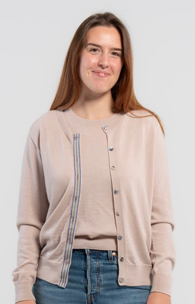 Woman wearing Noyac Knits Isabelle Classic Cardigan in light beige trimmed with signature grosgrain ribbon in stripes of tan, purple, lavender, and teal and Japanese mussel shell buttons -- knit in lightweight premium merino -- styled with matching merino wool crew neck sleeveless shell, jeans, ballet flats on plain background