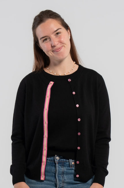 Woman wearing Noyac Knits Kate Classic Cardigan in black premium lightweight merino and trimmed with signature pink buttons and hot pink, light pink and green striped grosgrain ribbon trim styled over matching black merino crew neck sleeveless sweater shell and jeans on plain background.