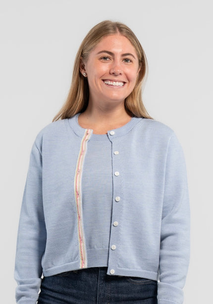 Woman wearing Noyac Knits Mary crop cardigan in soft, premium merino and trimmed with white mother of pearl buttons and signature ribbon in stripes of pastel pink, blue and green striped grosgrain ribbon styled with jeans on plain background.