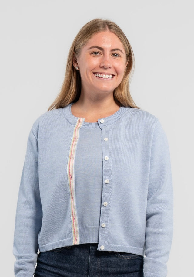 Woman wearing Noyac Knits Mary crop cardigan in soft, premium merino and trimmed with white mother of pearl buttons and signature ribbon in stripes of pastel pink, blue and green striped grosgrain ribbon styled with jeans on plain background.