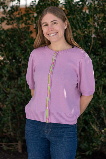 Woman wearing Noyac Knits lilac pink short puff-sleeve cardigan with pink and green ribbon trim, in premium, lightweight merino, styled with jeans against green leafy background