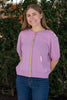 Woman wearing Noyac Knits lilac pink short puff-sleeve cardigan with pink and green ribbon trim, in premium, lightweight merino, styled with jeans against green leafy background
