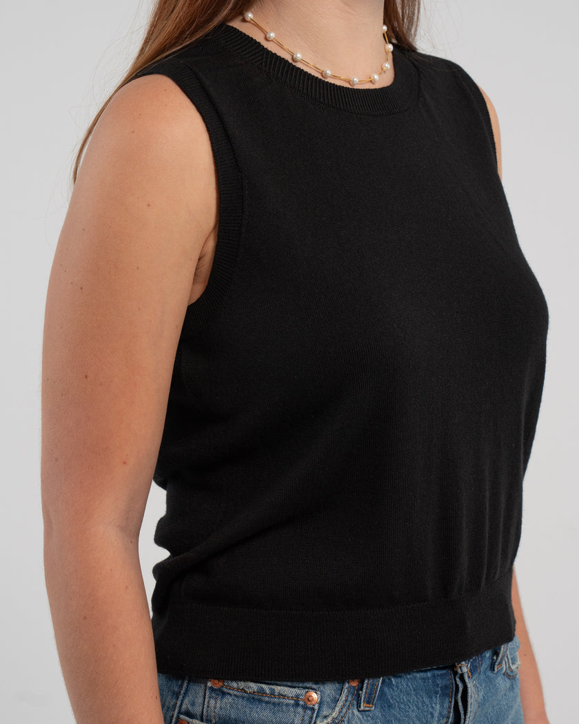  Close up of woman wearing Noyac Knits classic sleeveless sweater shell in black knit in ultra soft premium lightweight merino styled with jeans, pearl choker on plain background.  This versatile piece works well alone, over a blouse, under a matching classic cardigan or under a blazer.  