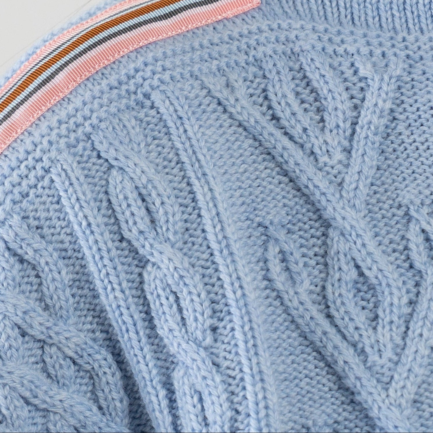 Close-up Noyac Knits Channing boatneck pullover in beautiful baby blue featuring cabling and signature ribbon trim in stripes of pink, white, light and dark blue and brown on shoulder, knit in premium soft merino wool  on plain background.