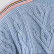 Close-up Noyac Knits Channing boatneck pullover in beautiful baby blue featuring cabling and signature ribbon trim in stripes of pink, white, light and dark blue and brown on shoulder, knit in premium soft merino wool  on plain background.