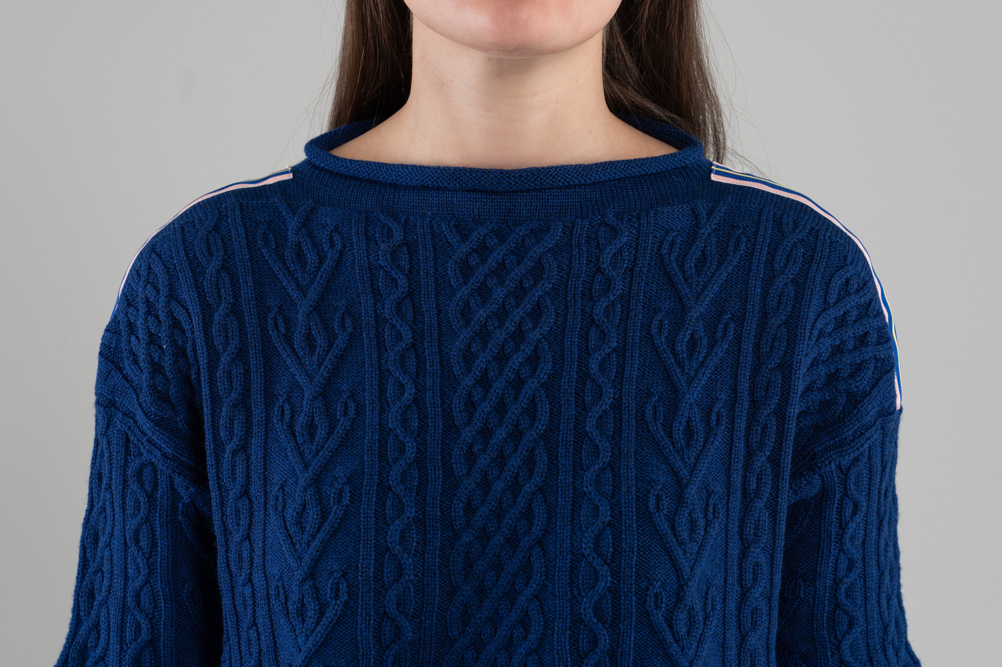 Close up of Noyac Knits cobalt blue boatneck pullover sweater with gorgeous cable detailing and signature ribbon trim in stripes of blue, pink and yellow on shoulder, knit in premium, soft merino wool  and plain background.