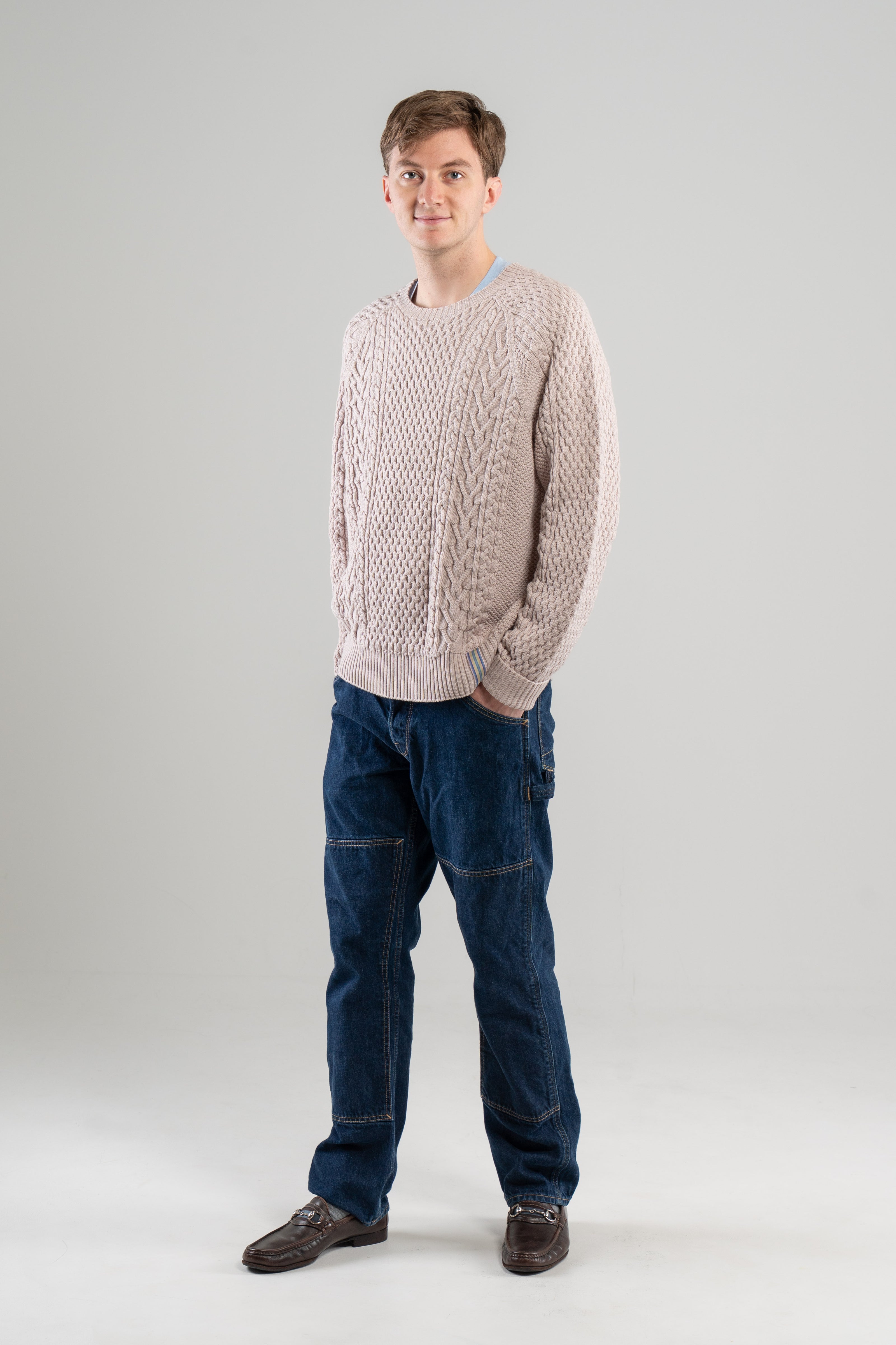 Man wearing Noyac Knits Cooper beige cable-knit sweater with signature ribbon trim on hem -- knit in premium merino wool -- styled with  jeans and loafers on plain background.