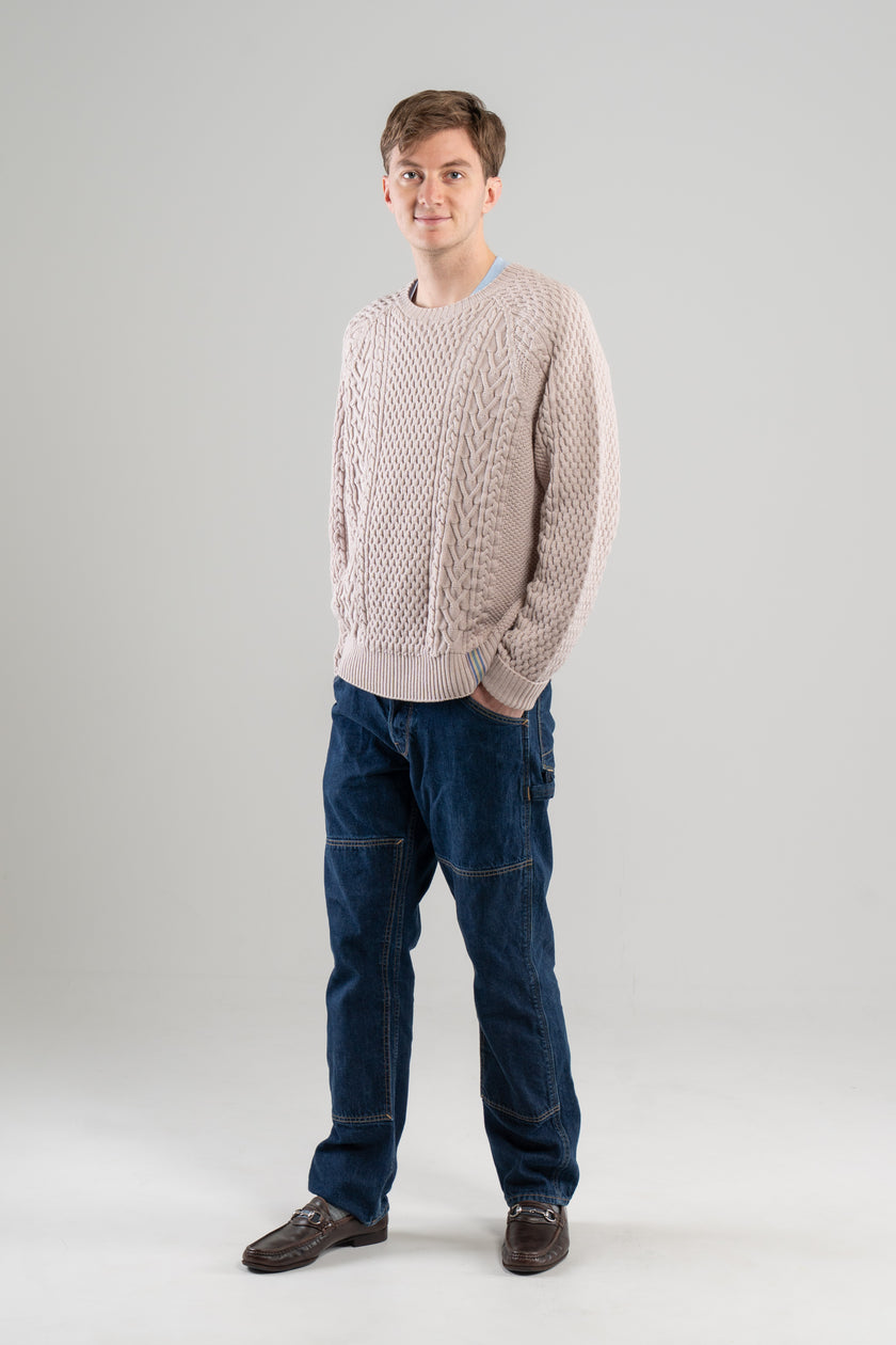 Man wearing Noyac Knits Cooper beige cable-knit sweater with signature ribbon trim on hem -- knit in premium merino wool -- styled with  jeans and loafers on plain background.