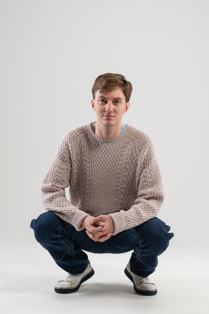 Man wearing Noyac Knits Cooper cableknit sweater in beige with signature ribbon trim on hem -- knit in premium merino wool -- styled with  jeans and sneakers on plain background.