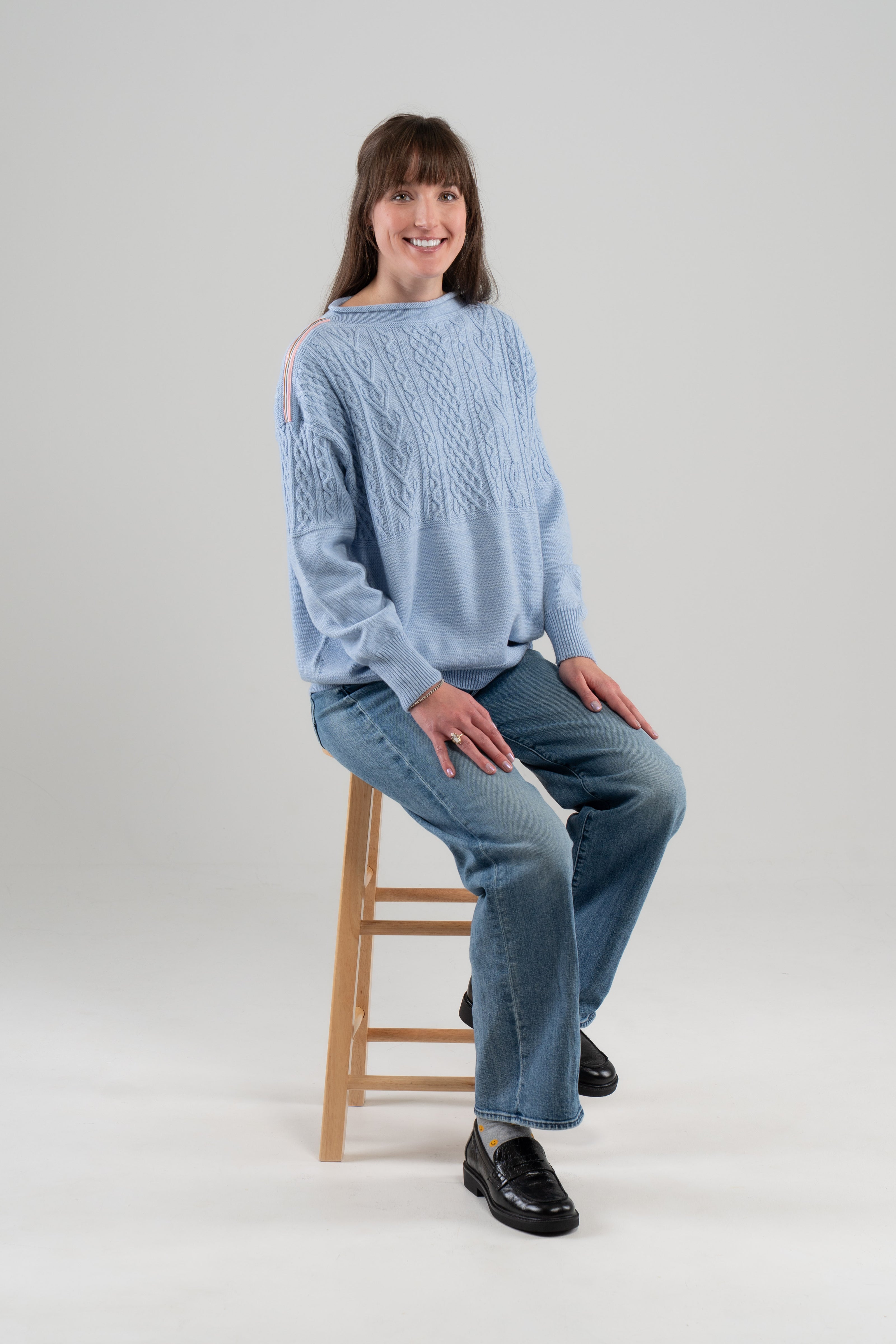 Woman wearing Noyac Knits Channing boatneck pullover in beautiful baby blue featuring cabling and signature ribbon trim on shoulder, knit in premium soft merino wool and styled with  jeans and loafers seated on stool on plain background.