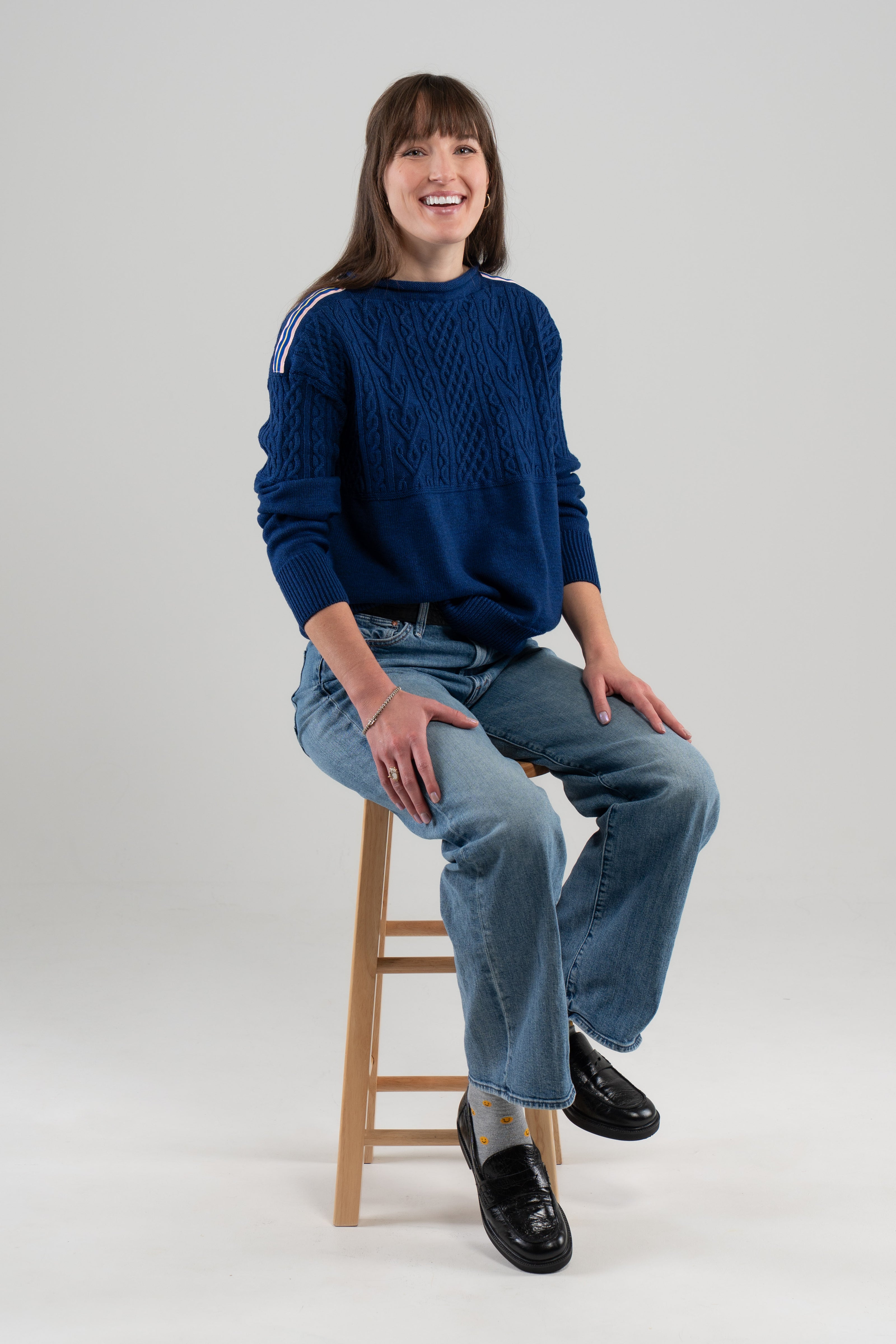 Woman wearing Noyac Knits cobalt blue boatneck pullover sweater with gorgeous cable detailing and signature ribbon trim in stripes of blue, pink and yellow on shoulder, knit in premium, soft merino wool and styled with jeans and loafers sitting on stool and plain background.