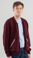 Man wearing Noyac Knits bomber-style sweater in burgundy color with signature striped ribbon-trimmed pockets -- knit in premium, ultrasoft merino wool -- styoed with  light blue t-shirt and jeans on plain background.