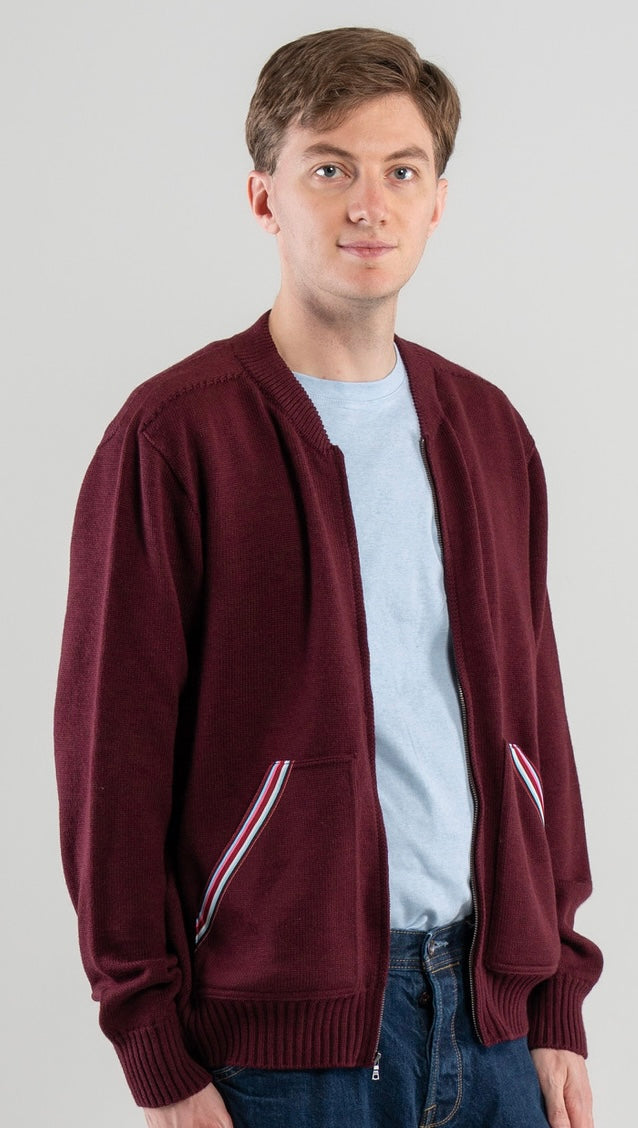 Man wearing Noyac Knits bomber-style sweater in burgundy color with signature striped ribbon-trimmed pockets -- knit in premium, ultrasoft merino wool -- styoed with  light blue t-shirt and jeans on plain background.