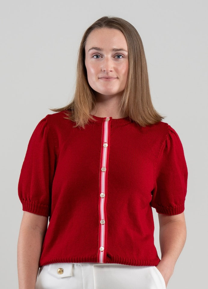 Woman wearing red Noyac Knits Rhoda short, puff-sleeve cardigan with signature ribbon trim in red, pink and white stripes   and white mother of pearl buttons, knit is soft, lightweight merino and styled with white pants on plain background.